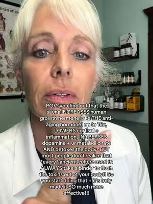 A medium close-up shot features a woman with short, light-colored hair looking directly at the camera. She has fair skin and is wearing a white lab coat. The background is a room with a framed anatomical chart, shelves with various bottles and containers, and a window.