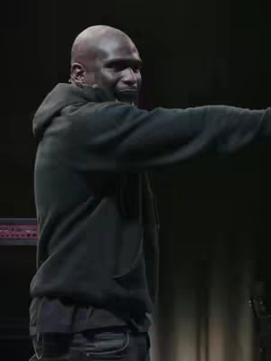 A medium shot shows a dark-skinned man with a bald head, wearing a black hooded sweatshirt and dark jeans, on a stage. His right arm is extended to the right, as if gesturing. The background is dark, with soft lighting emphasizing the man.
