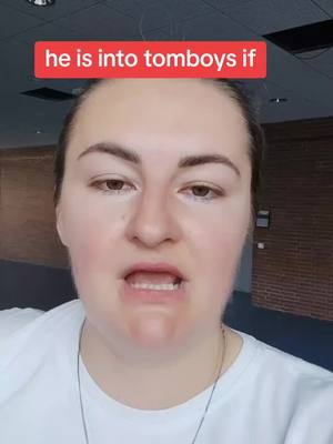 A selfie-style shot featuring a woman addressing the camera with a neutral expression. Above her, there is a text overlay that reads 'he is into tomboys if' within a red rectangular box. The background appears to be an interior space.