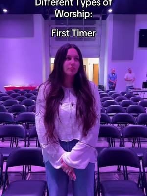 The image is a medium shot taken inside a church setting with a woman standing in the foreground. She is centered in the frame, wearing a light-colored, long-sleeved shirt and jeans. Her hair is long and dark, and she is looking straight ahead with a neutral expression. The background shows rows of empty black chairs and two people standing behind the chairs. The lighting is dim, creating a purple hue.