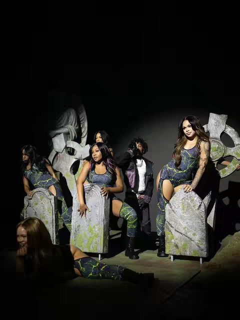 A group of diverse women pose in a theatrical environment. They are dressed in matching outfits with a marbled stone effect, with green accents. Each one is placed individually in front of tombstones, with a large angel statue in the background.