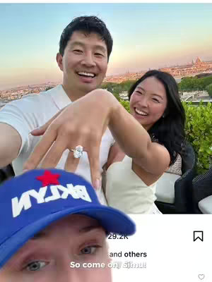 The image is a selfie-style shot featuring a smiling Asian man, Simu Liu, in the foreground, holding up a hand to display a large engagement ring. Behind him is a smiling woman, assumed to be his fiancee, and in the background, there's a blurred cityscape, possibly Paris, with warm lighting suggesting either sunrise or sunset. A blue baseball cap with white text is in the lower portion of the image, with a fragment of text from a social media app overlaid near the bottom, including the creator's eyes looking up.