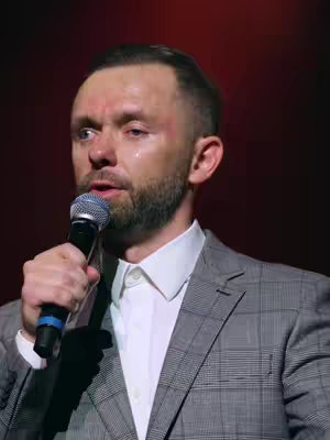 The image features a man with short, neatly styled brown hair and a trimmed beard. He is wearing a white dress shirt and a gray plaid blazer. The man is holding a microphone close to his mouth with his left hand, suggesting he is speaking or performing. His expression is serious, and there are apparent tear tracks coming from his eyes, possibly indicating emotional intensity. The background is a dark red, focusing attention on the man.