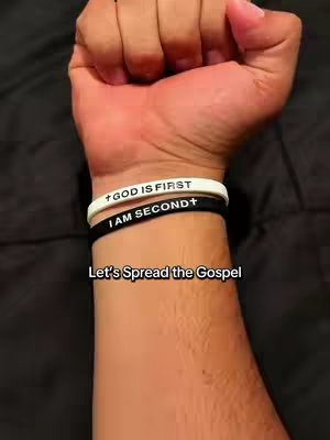 The image shows a person's arm with two wristbands. The top wristband is white and says 'GOD IS FIRST' in black text, and the bottom band is black, with the text 'I AM SECOND+' in white. Below the wrist appears to be a handwritten text: 'Let's Spread the Gospel'.