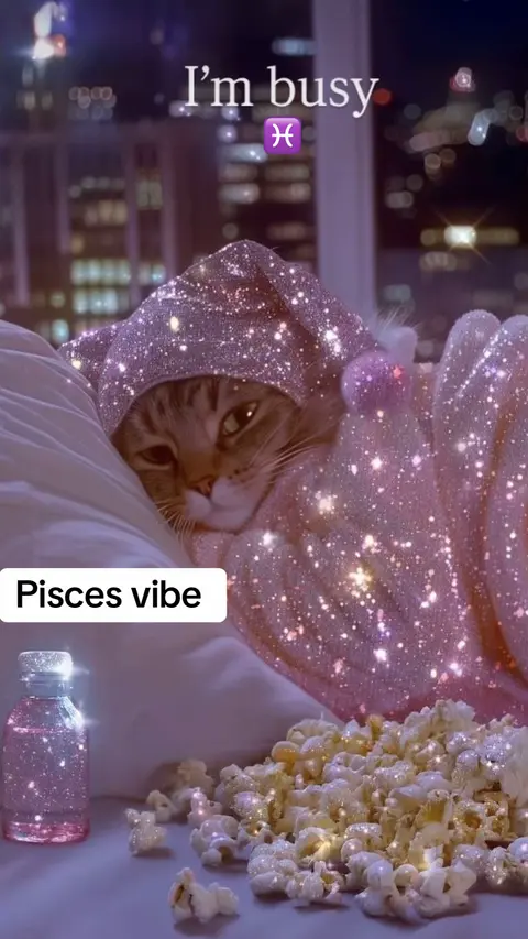 A close-up shot of a cat nestled in bed, wearing a sparkly pink hooded outfit and surrounded by pillows. Overlayed text reads 'I'm busy' and 'Pisces vibe.'  Additional elements include a small bottle filled with pink liquid and a scattering of popcorn, both also embellished with sparkles.