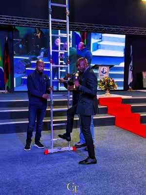 A group of men on a stage, with a ladder positioned upright in the center. One man is climbing the ladder while the others are standing on the stage below, wearing various outfits. The background displays a stage setting with screens and a podium with a logo.