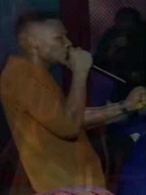 A man with short hair speaks into a microphone, likely in a stage setting. He wears an orange shirt, and is positioned to the left. The background is blurry but includes other people and some blue and red color.