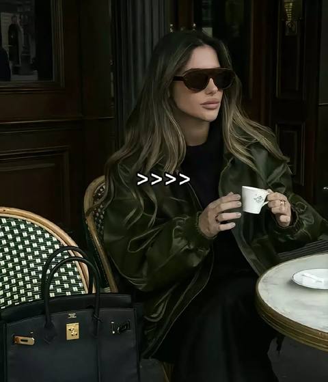A woman in sunglasses sits at a cafe, holding a cup of coffee. She's well-dressed and the setting is elegant.