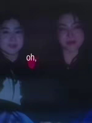Two young women are visible in a dark scene, filmed from the chest up. The women have dark hair and are positioned close together. The upper left corner has text that says "oh,". The background is dark with hints of blue light.