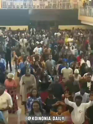 An overhead shot of a large crowd gathered in an indoor auditorium. Most people are dressed in casual attire, and many are raising their hands in the air. The room is brightly lit, and the focus is on the multitude of individuals participating in the event. Text overlayed on the image reads "@KOINONIA DAILY".