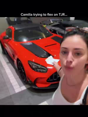 A woman is standing in front of a red sports car and wiping it with a white cloth. The car is parked in what appears to be a garage or parking structure. The woman is wearing a white top and appears to be posing or filming.