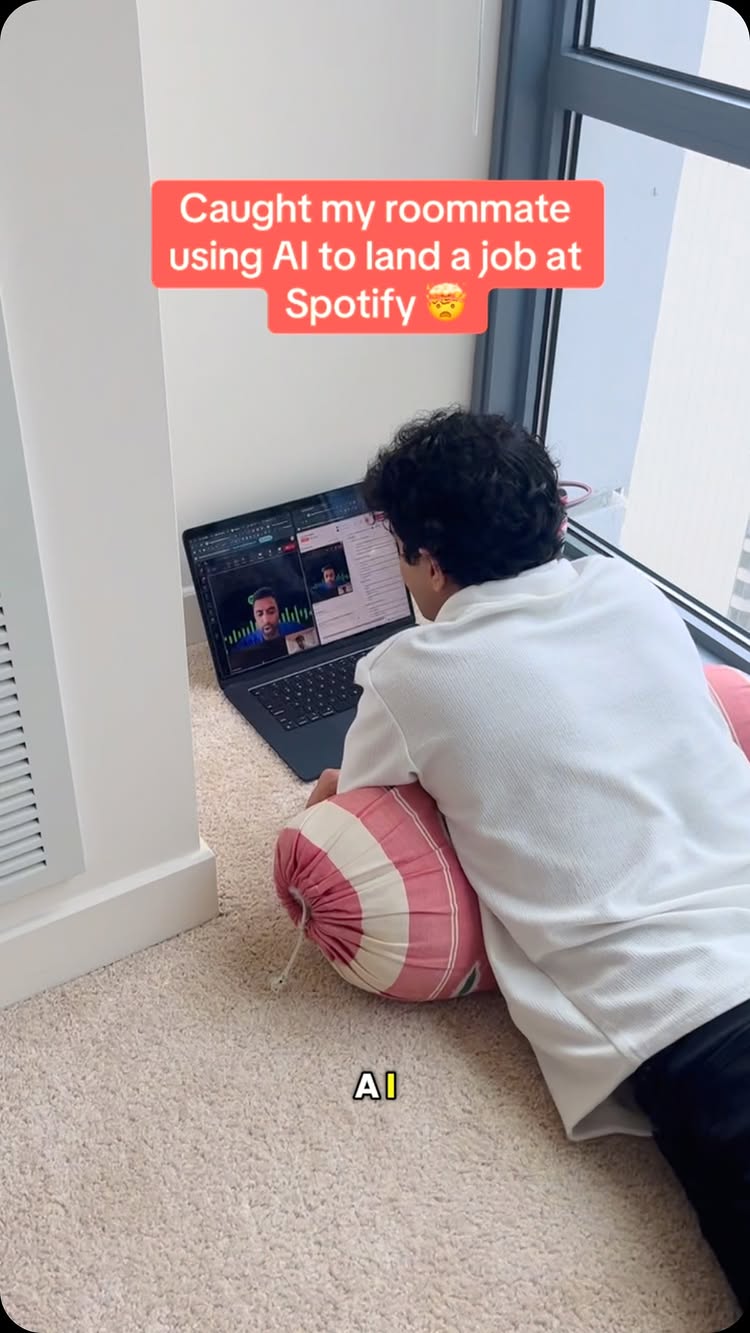 A person is sitting on the floor in front of a laptop, appearing to be in a video call. The scene is set in a room with a window and carpet. There is text overlayed on the image in a red box that reads, 'Caught my roommate using AI to land a job at Spotify'. The individual is wearing a white shirt and a colorful pink and red striped pillow.
