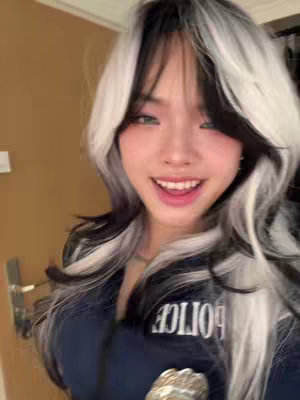 A close-up shot of a young woman in a cosplay outfit, smiling at the camera in front of a door, the background being out of focus. The woman has long black and white hair and is wearing a navy blue outfit with the word 'POLICE' visible on her chest. She has fair skin, pink lipstick and appears to be indoors, with golden-brown colored door in the background.