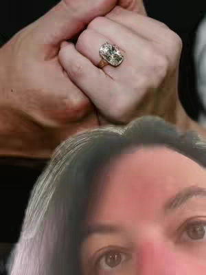 A close-up image featuring the hands of a person holding a large, sparkling oval-shaped diamond ring. The hand is delicately holding the ring up, the bottom half of the image has a person’s eyes and hair in the background