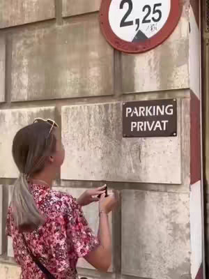 A woman with sunglasses on her head reaches towards a wall, likely interacting with something mounted on it. The setting appears to be outdoors with a concrete wall background and a sign indicating either a parking restriction or some sort of specific height clearance.