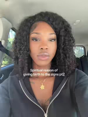 A medium shot of a woman with dark curly hair, wearing a black zip-up hoodie and a gold flower necklace, sitting in a car. The background shows the car's interior and a glimpse of trees outside the window. The overall tone is casual and conversational.
