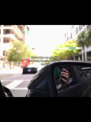 The image shows a view from inside a car, looking out through the side mirror. The primary focus is on the reflection in the mirror, where a person is holding a phone up to their face, presumably taking a photo or video. The background includes a city street with buildings on both sides, other cars, and traffic lights, suggesting an urban setting.