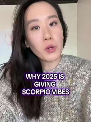 A medium shot of a woman with long dark hair, wearing a gray sweater, looking directly at the camera. The image has a text overlay in a purple box with the caption 'WHY 2025 IS GIVING SCORPIO VIBES'. The background is a light color, likely a wall, with a hint of something white in the left corner.