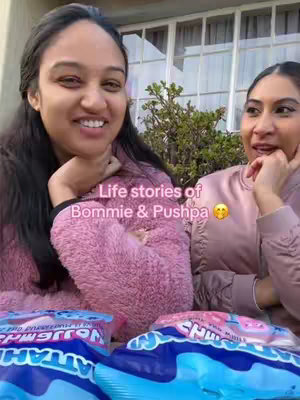 Two women are seated outdoors, one in a pink fuzzy sweater and the other in a pink top. They are smiling towards the camera. In the foreground, there are bags of Utz brand chips. Text overlay reads "Life stories of Bommie & Pushpa".