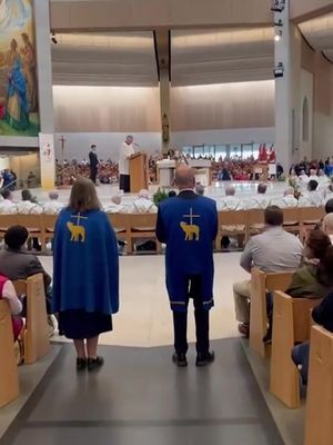 A wide shot of a large church interior at a Catholic ceremony.  Two people, seen from the back, are centered in the foreground. They wear matching dark blue capes or vestments which have a gold Lamb of God symbol on the back. Behind them, a man in a white robe is at a podium with a microphone, apparently delivering a sermon. Numerous people are sitting in the pews.