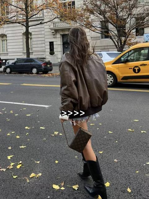 The image shows a person walking towards the viewer on a city street. They are wearing a brown bomber jacket, a denim skirt, and black boots. They are carrying a small, patterned handbag. The background includes a yellow taxi cab, buildings, and trees with autumn leaves.