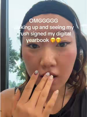 A close-up selfie shows a young woman with fair skin and dark hair pulled back in a headband. She's holding her hand up to her mouth, showing off detailed nail art. The background is slightly blurred, suggesting an indoor setting.