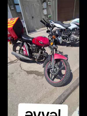 A motorcycle is parked on a concrete surface, with a large red storage container attached to the back. Another motorcycle is visible in the background, and the door and other parts of a building are in the background. The hook text is placed at the bottom.