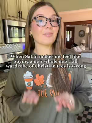 A woman with long brown hair and glasses is centered in the frame, wearing a sweatshirt with a coffee cup, leaves, and a football design. The background is a kitchen.