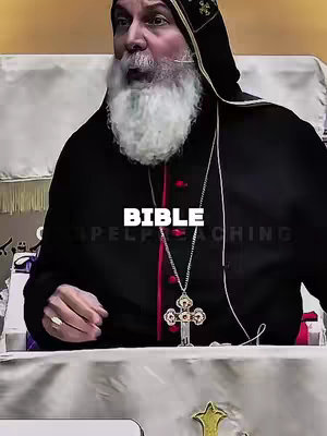 The image shows a medium shot of an older man with a white beard and dark attire, likely a religious figure such as a bishop or priest. He is positioned behind a table or pulpit, with his upper body visible, and he appears to be speaking or mid-sentence. A large silver cross hangs around his neck. The background appears to be an indoor setting, with a slightly off-white or cream-colored backdrop. Text overlays are positioned at the center. The overall composition is somewhat serious, given the subject matter and attire.