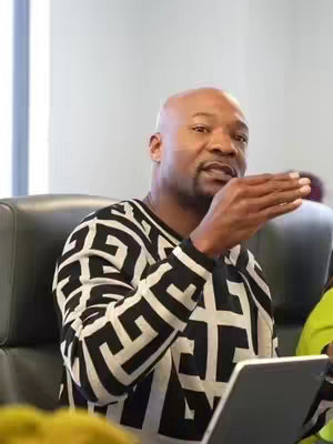 A medium shot features a bald Black man in his 40s or 50s, presenting an argument. He is wearing a black and white patterned sweater and his left hand is raised. His eyes are focused, suggesting an intent focus. The background suggests an office or studio setting, contributing to a professional ambiance. The color palette is mostly neutral with the strong black and white pattern providing the visual focus.