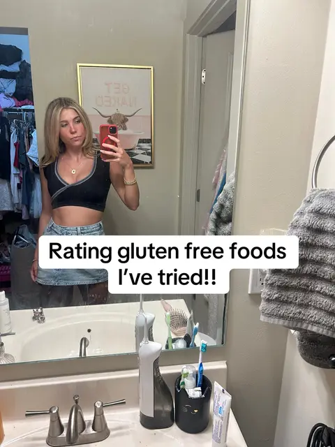 A woman in a bathroom takes a selfie in front of a mirror. She is wearing a black top and a denim skirt. The bathroom has a framed picture and various toiletries visible in the background.