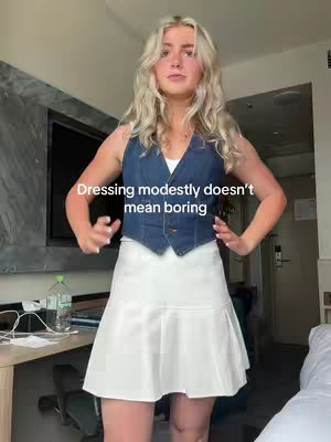 A young woman with long blonde hair stands facing the camera, hands on her hips, in front of a blurred background with a television and windows visible. She is wearing a denim vest over a white top and a white skirt. Text overlayed on the image reads: "Dressing modestly doesn't mean boring."