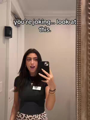 A woman is in a mirror selfie. The foreground shows the woman holding a phone in front of her face. She has long, dark hair, and is wearing a black top and a patterned skirt. Behind her is a bathroom; a door, a light fixture and part of framed mirror are visible.