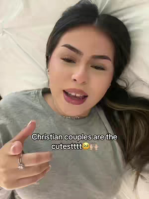 A young woman is lying on a bed, looking at the camera and speaking. She is wearing a gray shirt and has dark hair. Text overlay 'Christian couples are the cutest' is added to the image. The background is a white sheet.
