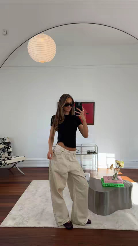A woman in a casual outfit is taking a mirror selfie in a well-lit room. She's wearing sunglasses, a black top, and wide-leg, beige pants. The composition includes a large mirror, a hanging circular light fixture, a chair, and a coffee table, suggesting an aesthetically pleasing interior.