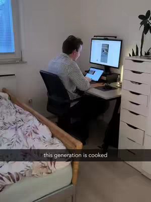 A man is sitting at a desk in a room, working on a computer. The room is sparsely furnished with a bed and a dresser visible. Text overlay reads 'this generation is cooked'.