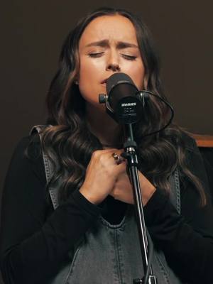 A woman with long wavy brown hair is positioned in front of a microphone. Her face is focused, eyes closed, and hands clasped in front of her chest. She's wearing a black shirt under a denim overall top. The background is dark, with a microphone stand and part of a wooden structure.
