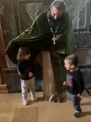 A priest in a green robe is standing amongst two children in a stone-colored room. The priest is looking down, seemingly interacting with the children. The children are looking at each other with playful expressions.