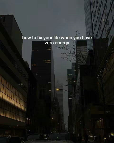An urban cityscape at dusk. Tall, dark buildings line both sides of a street, converging towards a point in the distance. The sky is a muted gray, and streetlights cast a warm glow. Added text overlay says 'how to fix your life when you have zero energy'.