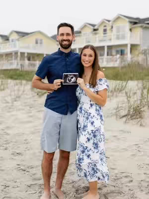 A couple stands on a sandy beach holding an ultrasound photo. The woman is wearing an off-the-shoulder white dress with blue floral patterns. The man is wearing a blue button-up shirt and gray shorts. They are both smiling. Behind them, the background is blurred, with a row of buildings visible.