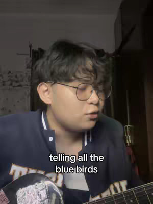 A young man with short, dark hair and glasses is singing, holding a guitar. The background is dim, suggesting a room. Text overlayed on the image reads lyrics.