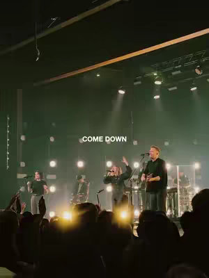 A live concert scene with a band on stage, illuminated by stage lights and spotlights. The crowd is partially visible in the foreground, with the band members in the middle ground. Text overlay 'COME DOWN' is visible at the top.