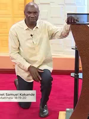 A man wearing a light-colored button-down shirt and dark pants kneels on a red carpet, with his right hand placed on a brown podium and his left hand resting on his knee. The background features a blurred geometric design.