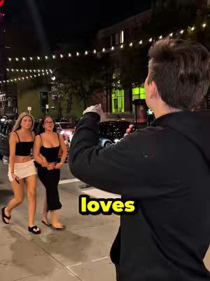 A shot taken at night, showing a man facing away from the camera, holding a phone and appearing to take a photo or video of two women walking on a sidewalk. The women walk side-by-side in the foreground. The background is blurred, but clearly indicates an urban setting with streetlights. Text overlayed on the image reads 'loves'.