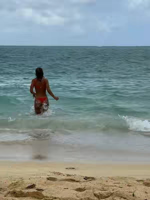 A woman in a red bikini walks from the ocean towards the sandy beach on a cloudy day. High angle, the water is a turquoise color.