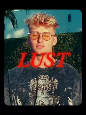A young man with blonde hair and glasses is centered in the frame. The word "LUST" in red text overlays him. He wears a dark t-shirt with a graphic design, set against a backdrop of green foliage and a light blue wall.