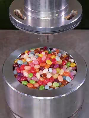 A close-up shot of a metal cylinder containing a variety of colorful jelly beans, possibly as part of a stress test by a solid metal press above.