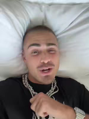 A man with short hair and a mustache is lying on a white pillow. He wears a black shirt with a decorative silver chain necklace and a silver watch on his wrist. He is looking at the camera.