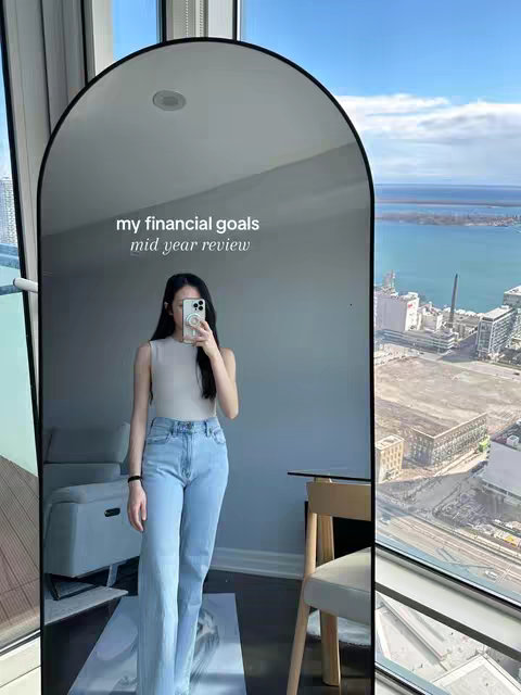 A woman is taking a mirror selfie in a high-rise apartment with a cityscape view. She's wearing a light beige sleeveless top and light-wash blue jeans. Text overlayed on the mirror says "my financial goals, mid year review". The mirror is framed by a black arch.