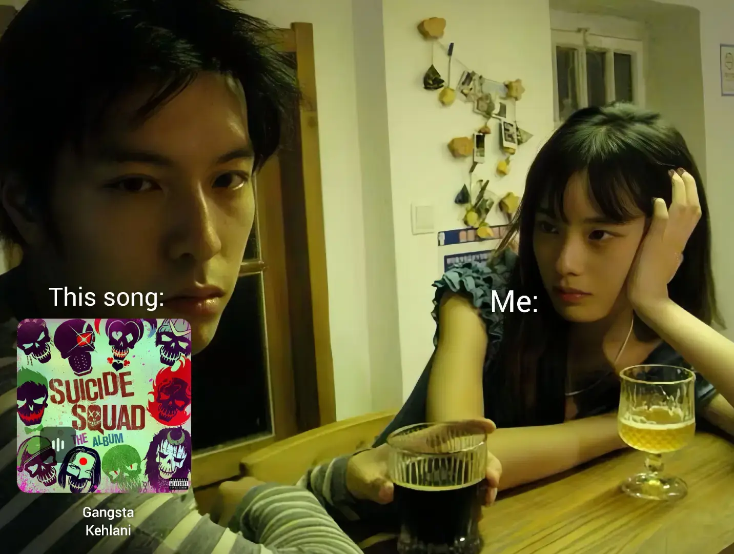 A scene in a dimly lit room with two people seated at a table. A man with dark hair looks directly at the camera with a serious expression, while a woman with dark hair, sitting to the right, has a more neutral expression with her hand on her face. On the table in front of the man is a beer and a Suicidal Squad album cover next to the text This song. Next to the woman is the text Me.